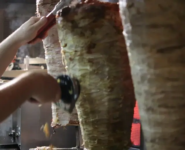 Three vertical spits of stacked meat, the left one being sliced with a handheld electric carver, a red-handled knife at the top of the middle one, a heating element glowing behind