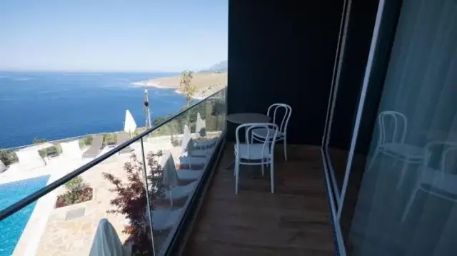 The view from a room's own balcony: bistro chairs on timber decking behind a glass balustrade, the pool and flagstone terrace below, and the coast running north far beneath