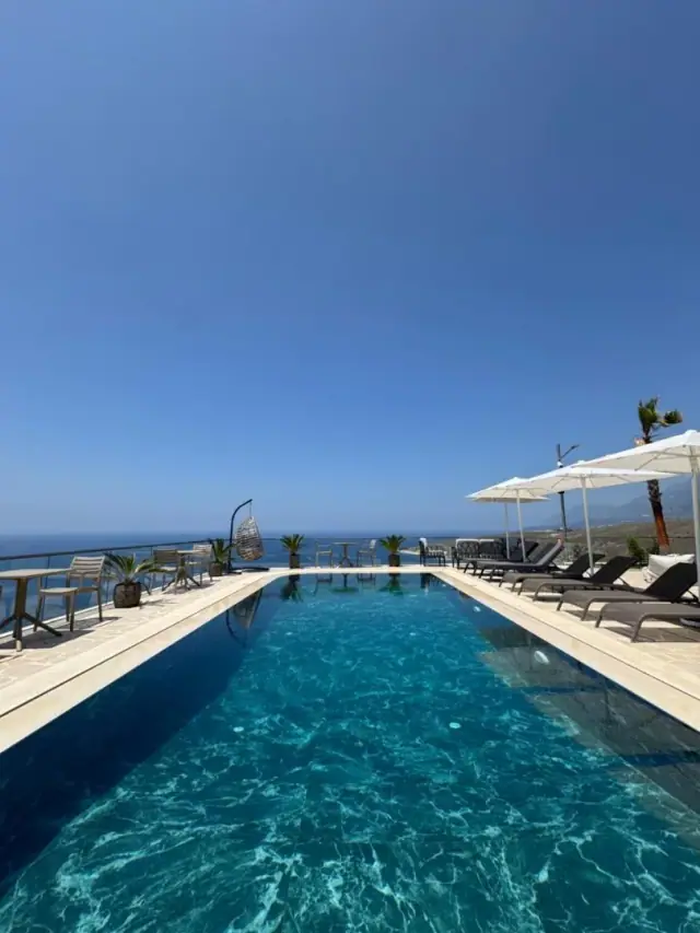The pool photographed from the water, its pale stone coping running out towards the Ionian far below, with white parasols, a hanging rattan chair and the coastline curving away to the right