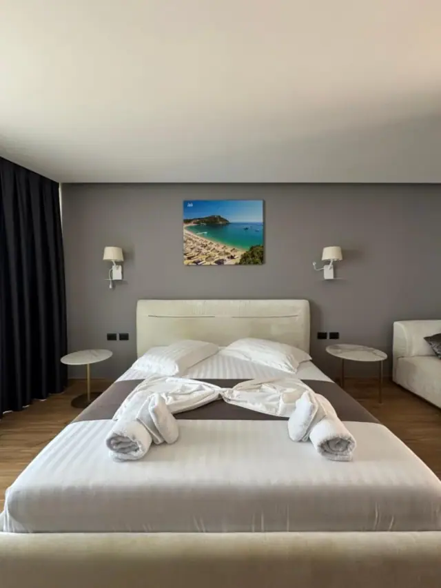 A made bed in striped white linen with rolled towels laid across it, marble-topped tables either side, and a framed photograph of a beach hung on the warm grey wall above the headboard