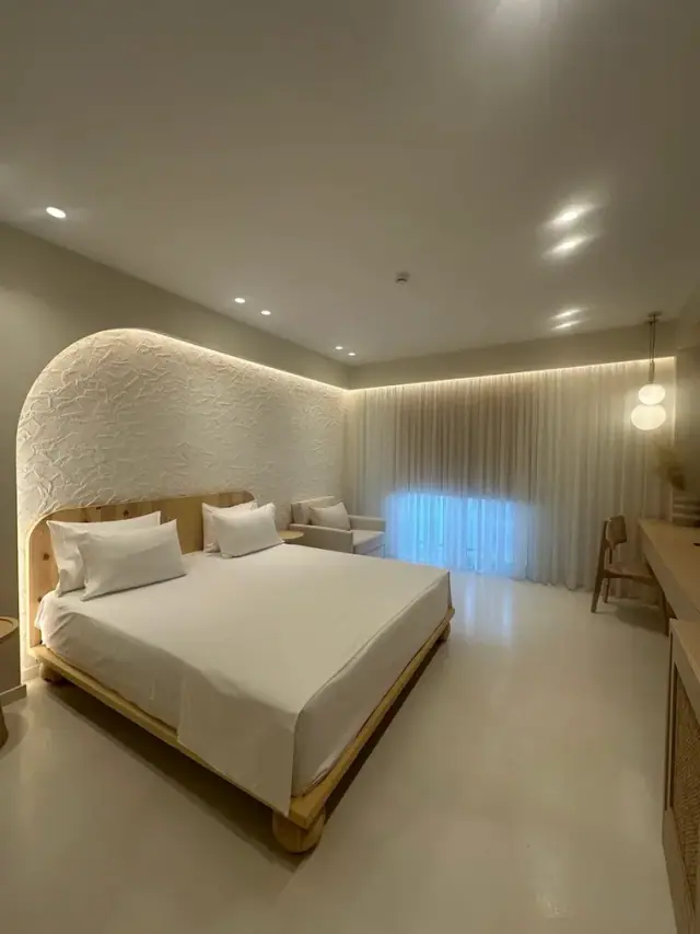 A room at Dimitri Hotel where the bed stands in a tall arched recess of textured white plaster, lit from behind, with a sheer curtain glowing across the far wall