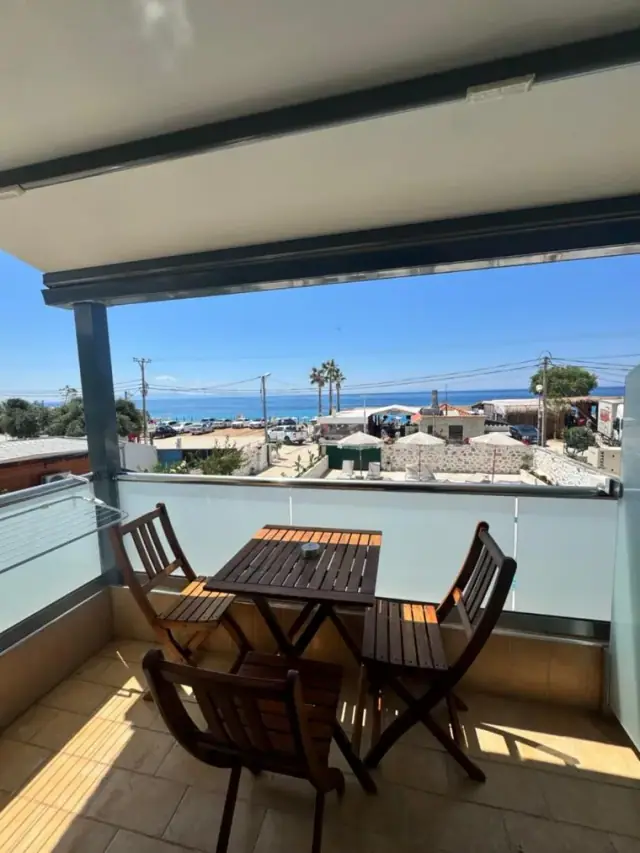 A shaded balcony at Dimitri Hotel with a slatted timber table and three folding chairs, looking past the pool parasols to the shingle and the Ionian