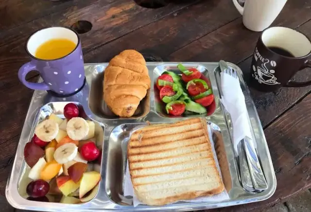 A breakfast tray at Himara Downtown Hostel on a plank table: fruit, a croissant, toasted bread, tomato and cucumber, with tea and coffee alongside