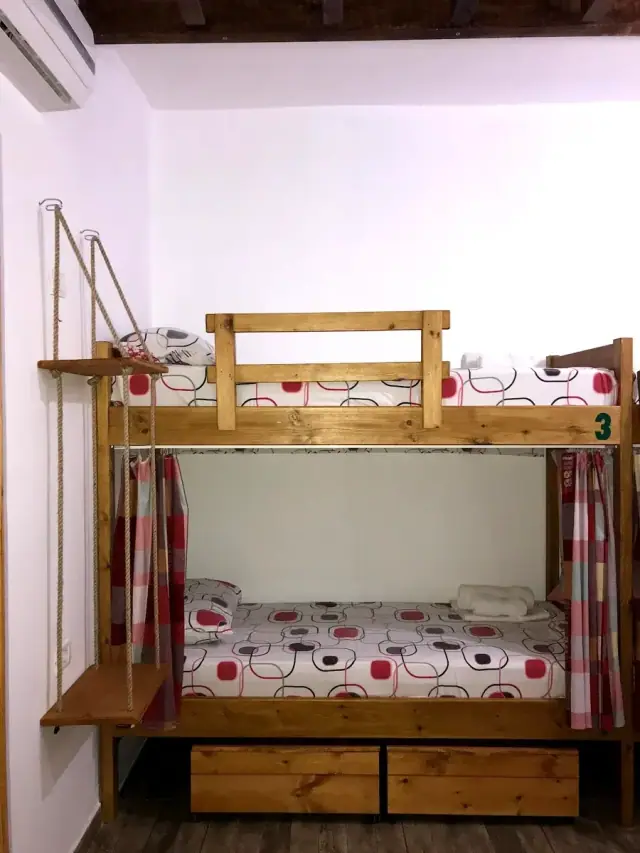 A pine bunk bed at Himara Downtown Hostel with a drawn privacy curtain, rope-hung shelves, drawers underneath and an air-conditioning unit above