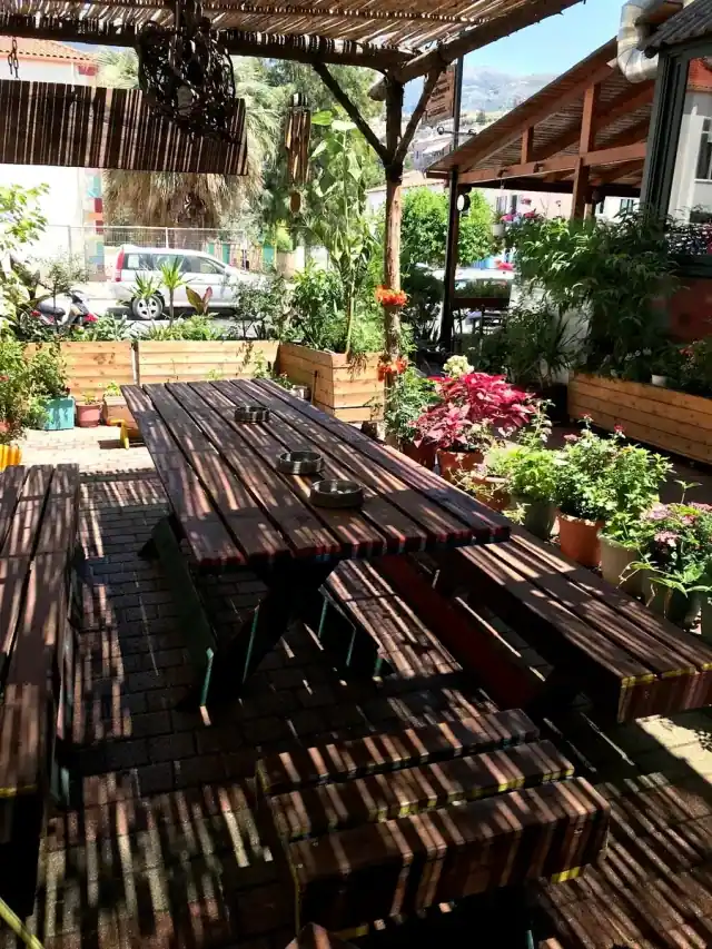 A long dark picnic table and benches under a reed pergola at Himara Downtown Hostel, with planters of flowers and the mountains behind