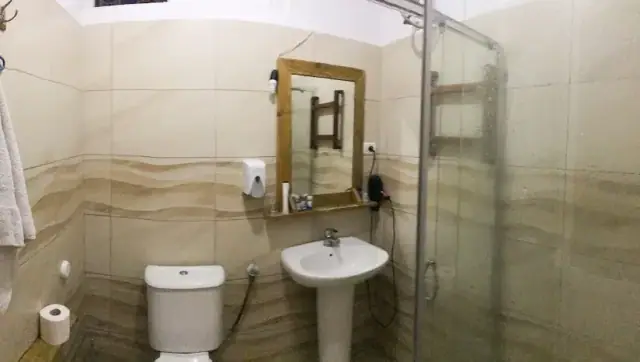 A shared bathroom at Himara Downtown Hostel with a tiled wall, a glass shower screen, a basin under a timber-framed mirror and a soap dispenser