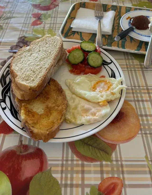 A breakfast plate of two fried eggs, fried bread, a slice of white bread, tomato and cucumber, on a fruit-patterned oilcloth beside a tray of jam