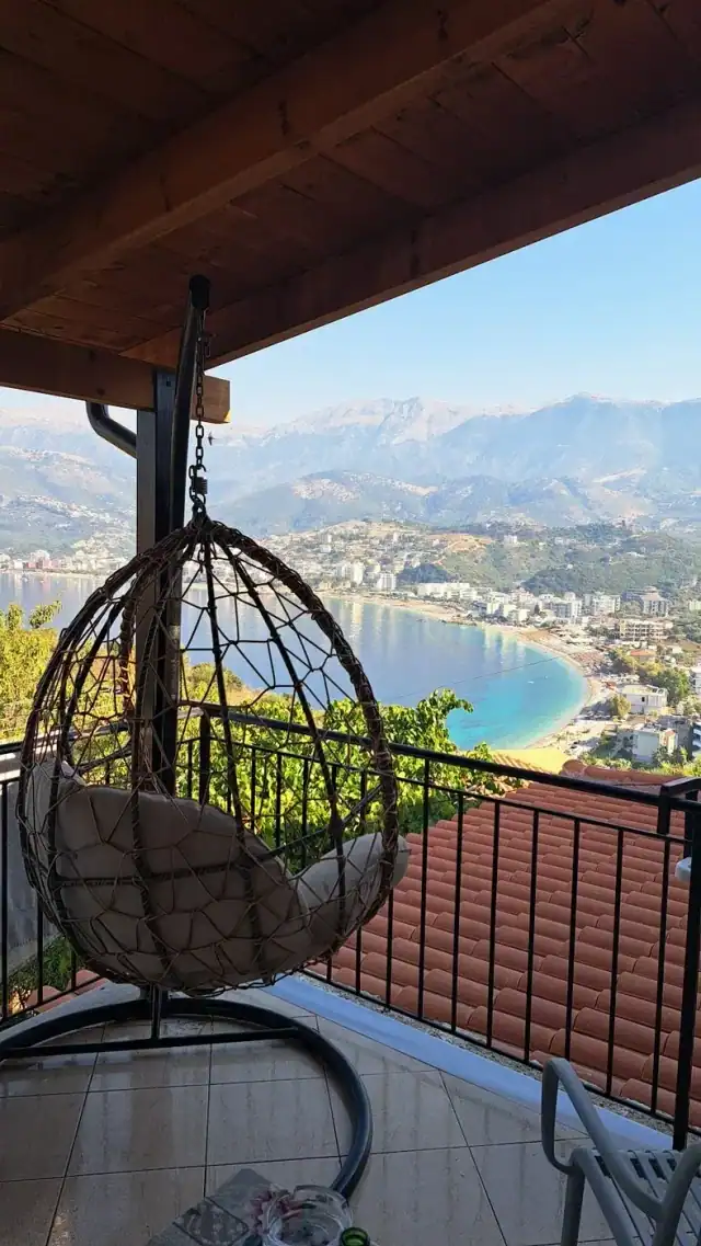 A wicker hanging chair on a tiled balcony under a timber roof, looking down over a curved bay, a town and the mountains behind it
