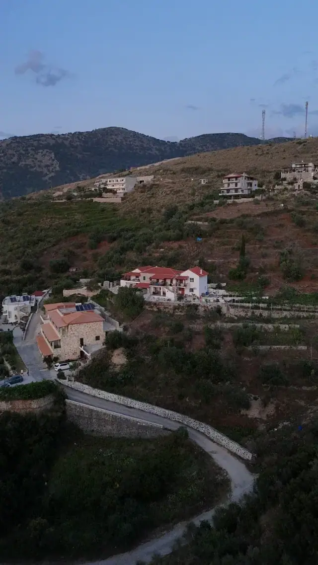 Aerial view at dusk of a narrow concrete road switching back up a hillside past stone houses and olive terraces