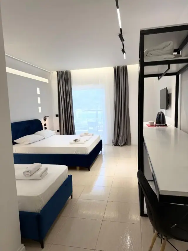 A room with a navy upholstered bed, grey curtains, a wall-mounted television and a worktop