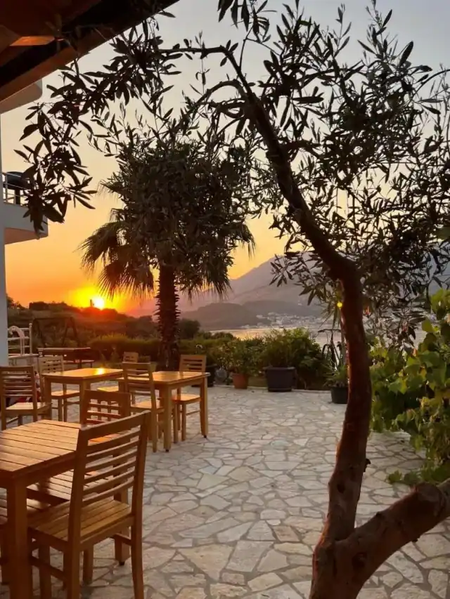 Wooden tables and chairs on a paved terrace under olives and a palm, the sun going down over the bay