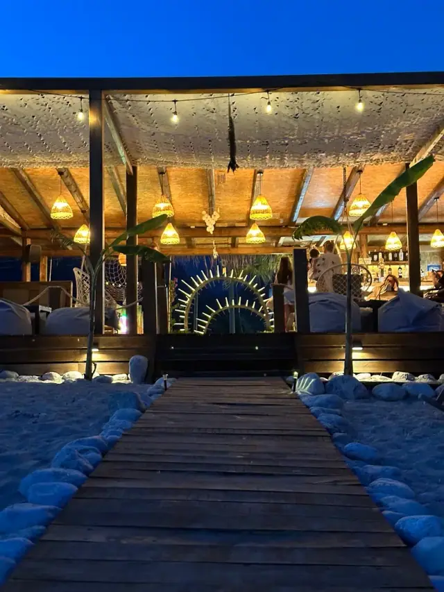 Boardwalk leading to the lantern-lit Paralian Beach Bar on the sand at dusk