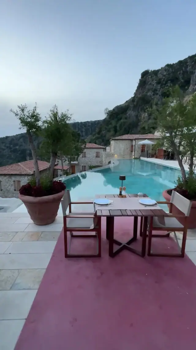 Infinity pool at Zoe Hora at dusk, flanked by stone village houses with tiled roofs and a limestone cliff behind, a set dinner table in the foreground