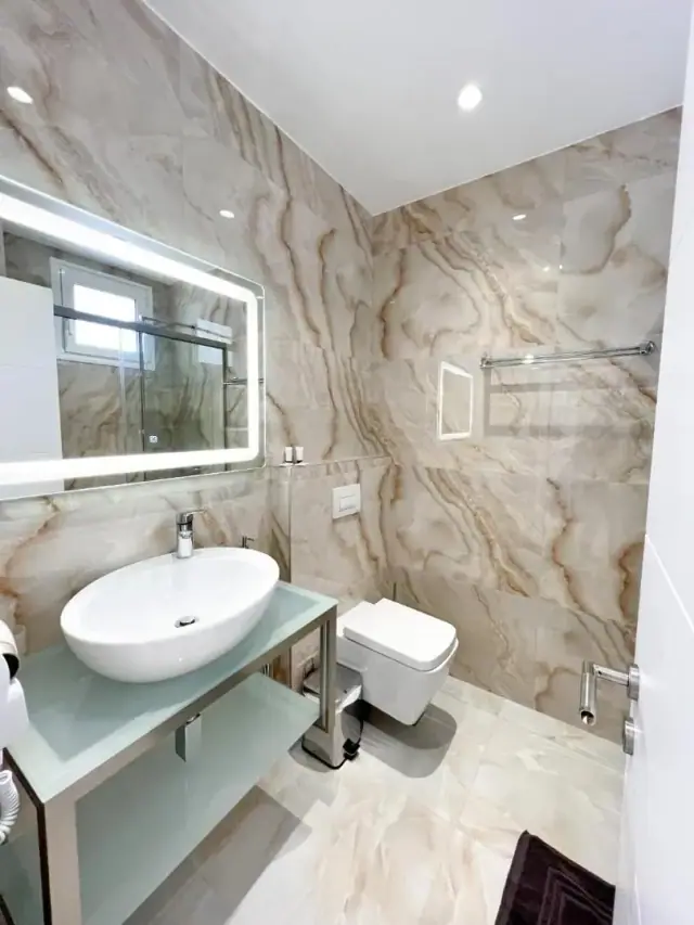 A bathroom lined in caramel onyx tile, with a vessel basin on a glass shelf and a lit mirror