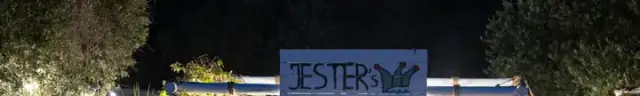 The hand-painted JESTER's sign, a blue plank board with the jester's-cap mark, standing above a rope-lashed white timber beam and lit at night
