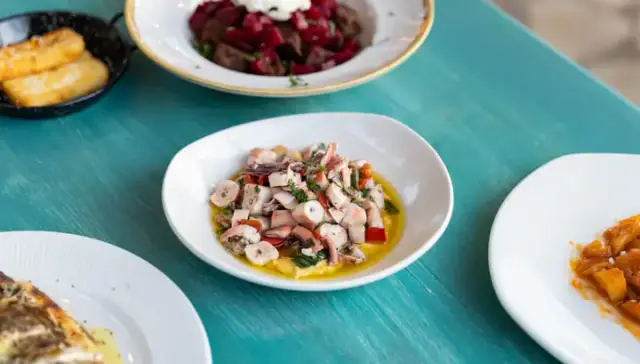 A white bowl of chopped octopus with herbs, red pepper and olive oil over a purée, on a turquoise painted table
