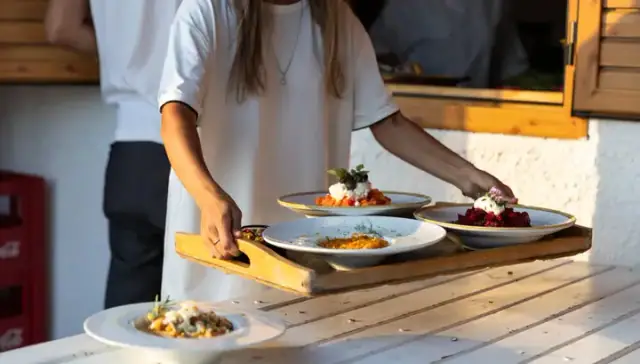 Plates of tomato salad, beetroot and a saffron-coloured dish carried out on a wooden tray to a white timber table in low sun