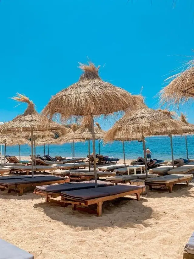 Straw parasols and wooden sun loungers on the sand at Prinos Beach, with the sea and a rocky point beyond