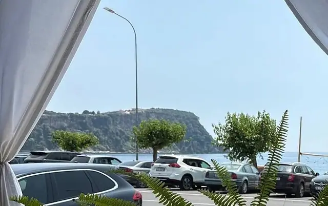 The view out from between the drapes: cars parked along the coast road, the water beyond them, and a headland standing across it.