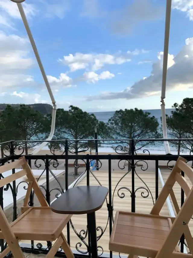 Two folding wooden chairs and a small table on a wrought-iron balcony, looking out over pines, the promenade and the sea at dusk