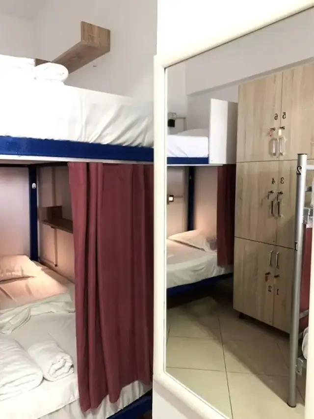 A bunk bed with its dark red curtain half drawn, folded towels on the mattress, and a column of numbered wooden lockers in the mirror beside it