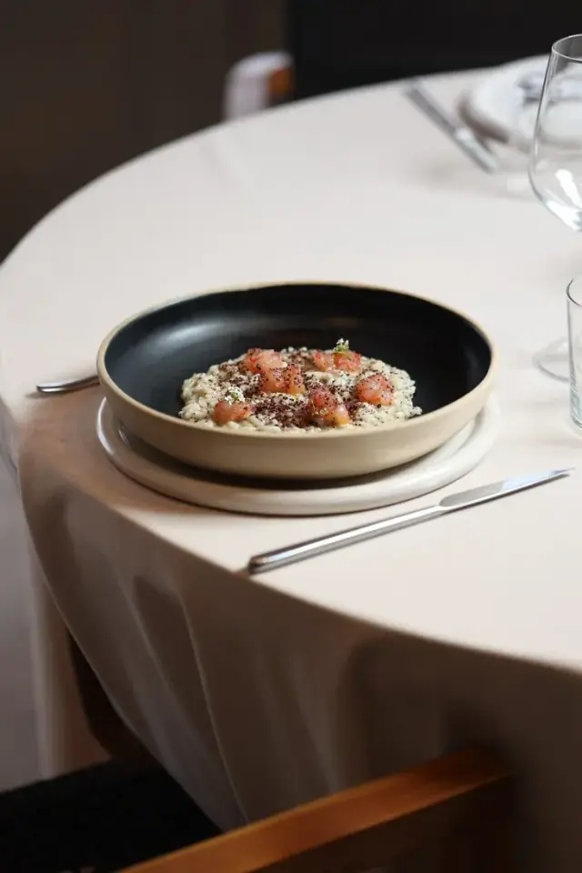A risotto finished with cured fish and herbs, in a dark bowl on a cream tablecloth