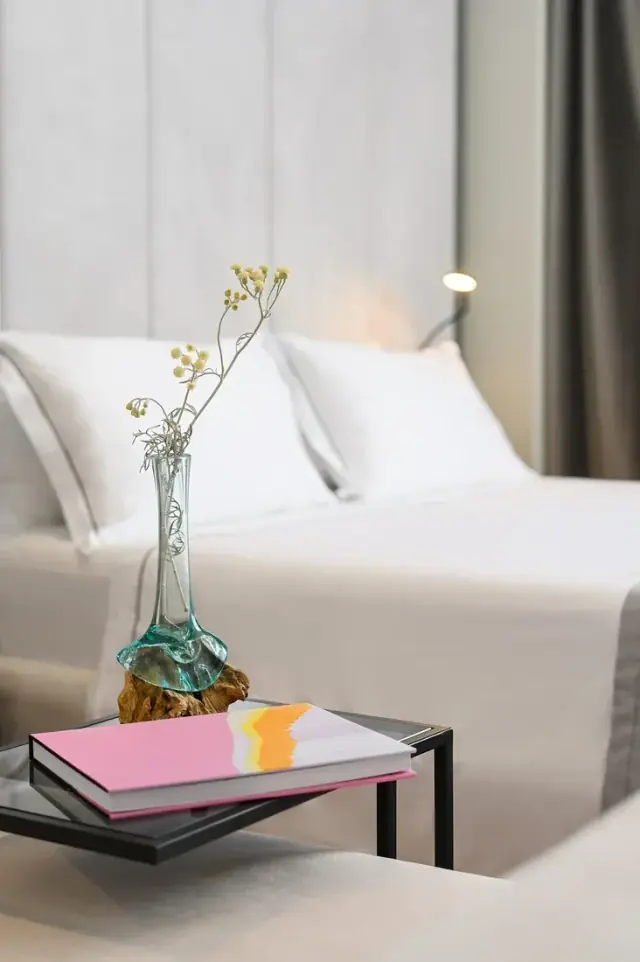 White bed linen with a hand-blown glass vase of dried flowers on a black side table