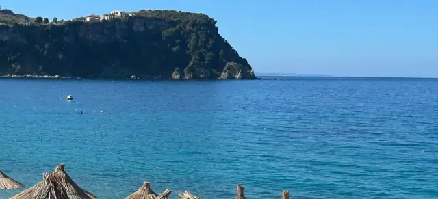 The wooded promontory that closes the south side of the bay at Namos Beach, with a small boat moored on flat turquoise water