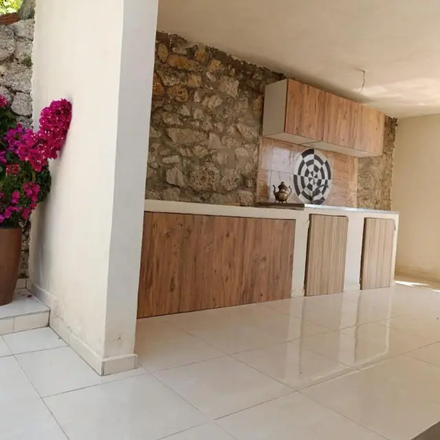 The outdoor kitchen at Nikolari Resorts: pale timber cupboards and a worktop built against a rough limestone wall, a brass pot on the counter and a bougainvillea in a terracotta jar at the corner