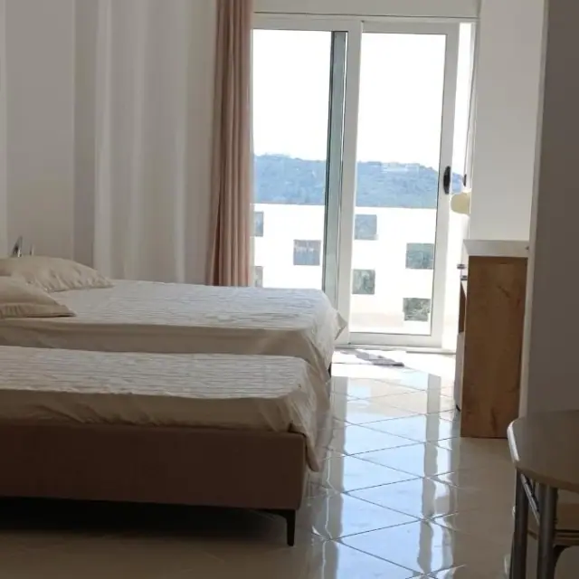Twin beds beside a sliding balcony door at Nikolari Resorts, looking out past a white parapet punched with square openings to a green ridge across the valley