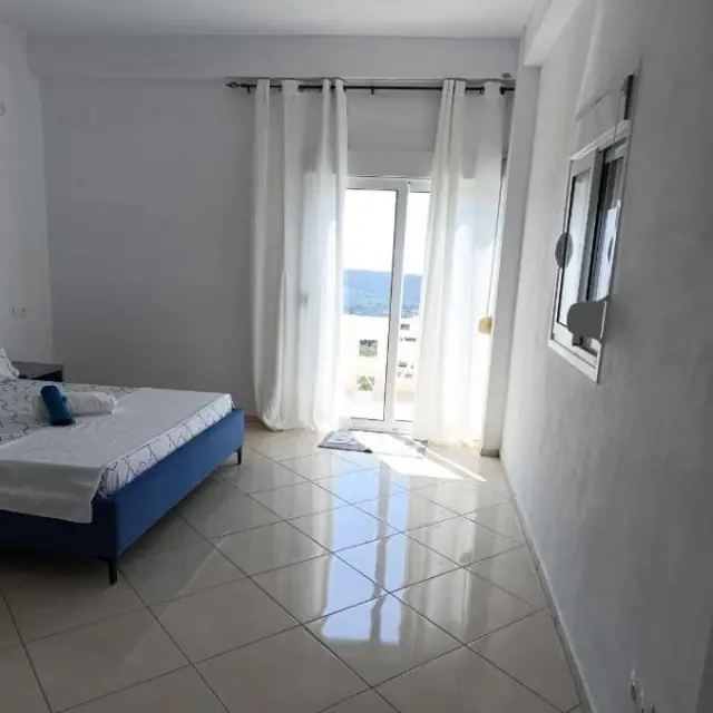 A bedroom at Nikolari Resorts with full-height white curtains drawn back from a balcony door, a framed mirror on the side wall and hills showing through the glass