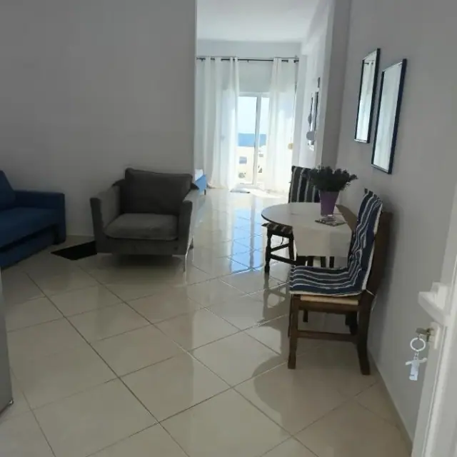 The sitting room of an apartment at Nikolari Resorts: a blue sofa, a grey armchair and a round dining table, with a pale band of water visible through the balcony door at the end