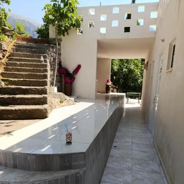 A long tiled counter running down a sunlit terrace at Nikolari Resorts, stone steps and magenta bougainvillea on the left, and a white concrete screen pierced with square openings spanning the passage above