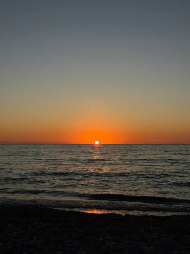 The sun touching a flat sea horizon off Spile beach, an orange band across the sky and a wet shingle foreshore in shadow