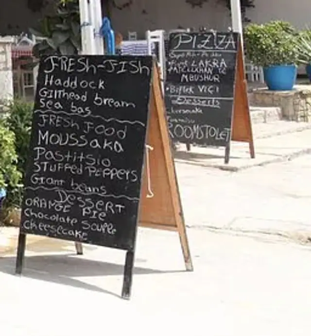 A handwritten chalkboard headed Fresh-Fish standing on the pavement at the road, with a second chalkboard headed Pizza further along