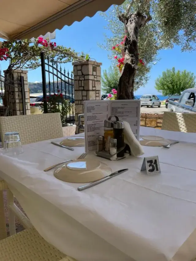 A table laid with white linen under a scalloped cream awning on the restaurant terrace, bougainvillea over the iron gate and an olive tree standing beyond the wall