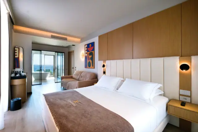 The bed in the Junior Suite with bath tub, sea beyond the balcony