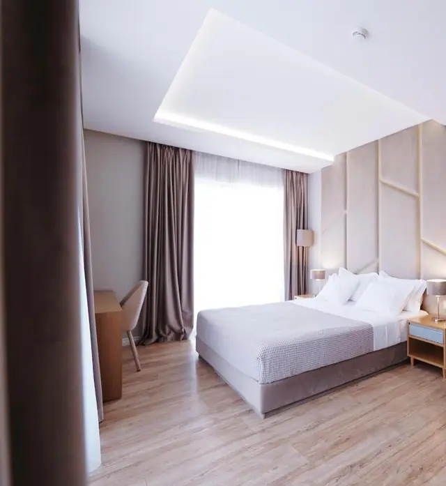 A hotel bedroom with a king bed against a wall of upholstered panels divided by slim brass battens, a matching table lamp either side, a recessed cove-lit ceiling, a writing desk and chair, and floor-length drapes over a full-height window