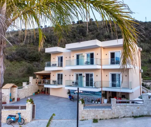 The seaward front of Seaside Hotel from its parking court, palm fronds overhead, a painted sea and the words Sea Side along the ground-floor wall