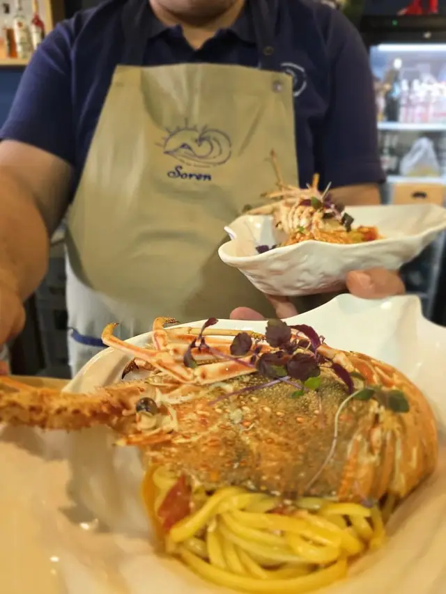 A whole spiny lobster set over long pasta on a white dish, carried out by a member of staff in a Soren apron