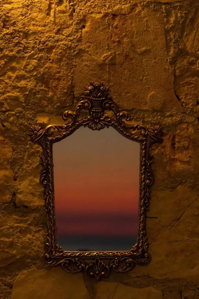 An ornate gilt-framed mirror hung on a stone wall, holding the reflection of the sunset