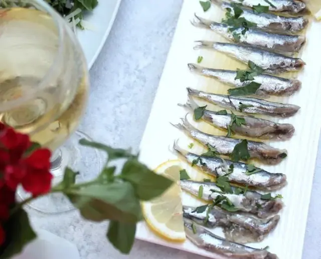 A long white plate of small silver fish, butterflied and dressed with olive oil, lemon and parsley, beside a glass of white wine