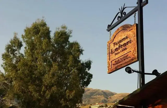 The carved sign of Taverna Lefteri hanging from a wrought-iron bracket, with the Himarë hills behind