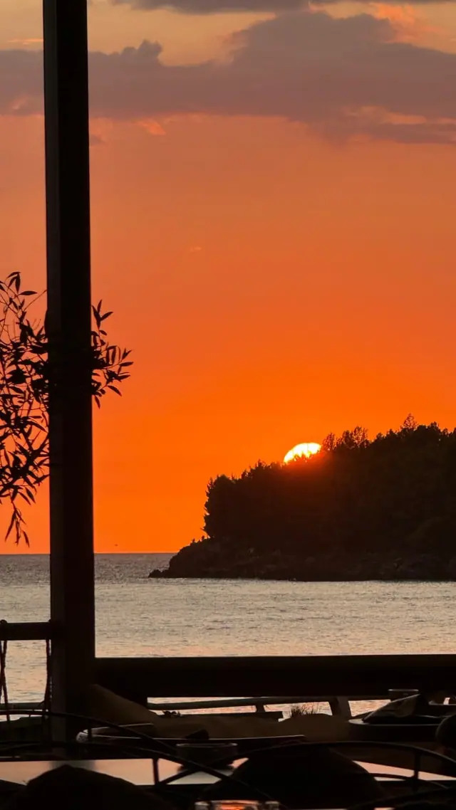 The sun dropping behind a wooded headland to the west-north-west, seen across the water on the Himarë shore