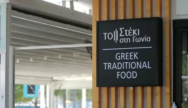 The black sign plate on the timber frontage, reading ΤΟ Στέκι στη Γωνία above the words Greek Traditional Food
