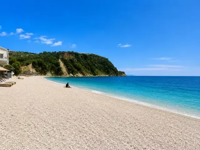 A wide white-pebble beach with turquoise water and a wooded headland at its far end