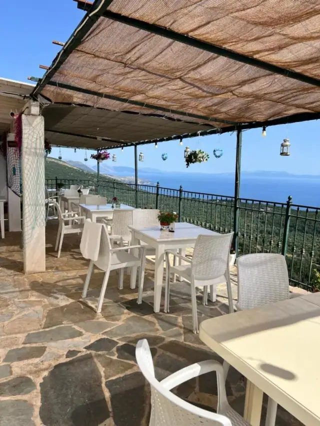 A terrace of white tables and chairs under a reed canopy hung with lanterns, the sea beyond the railing
