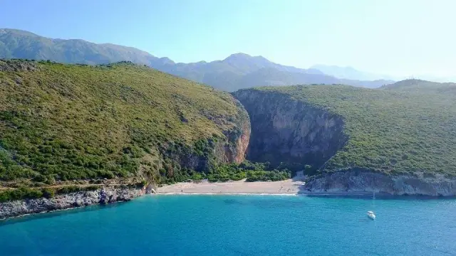 A cove of pale shingle between high cliffs, a boat anchored on turquoise water and a wooded gorge behind