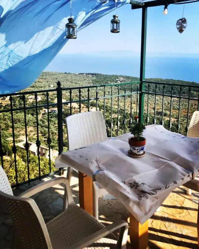 A laid table and a wicker chair on a balcony under a blue awning, olive terraces falling away behind