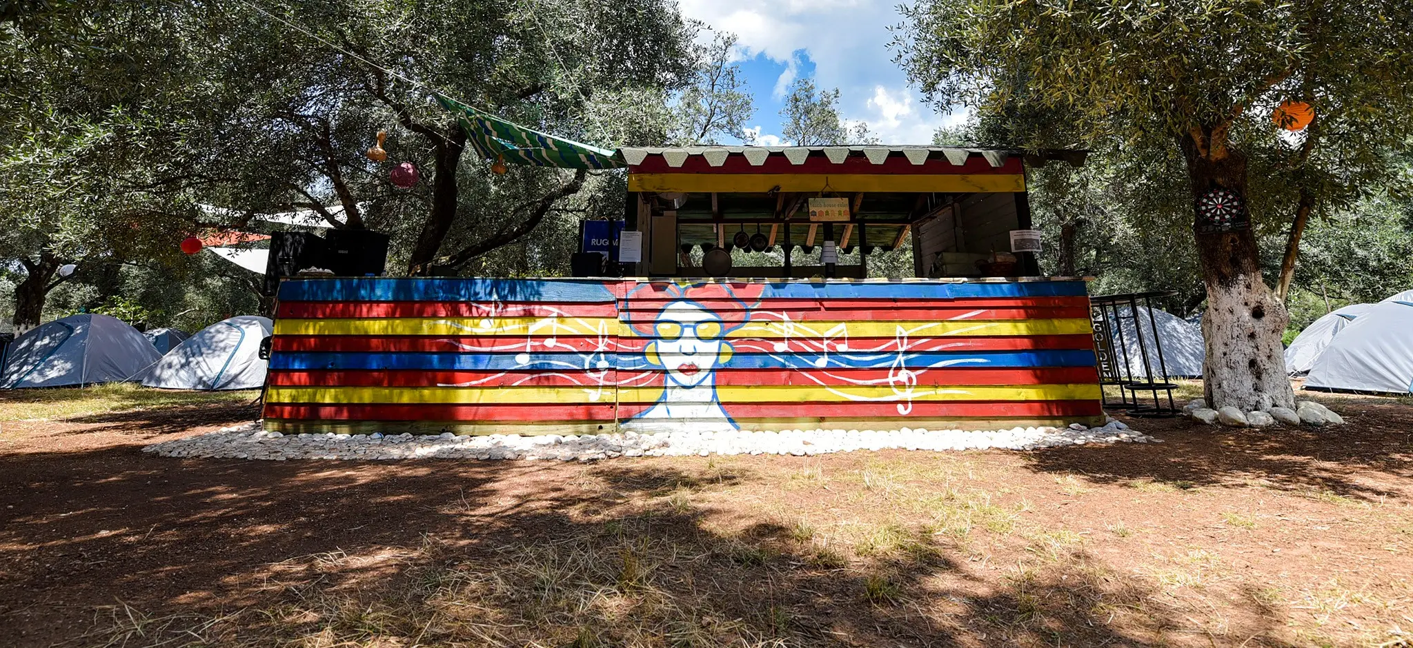 The camp's counter painted in blue, red and yellow planks with a mural of a woman in yellow sunglasses and music notes, tents under the olive trees on either side